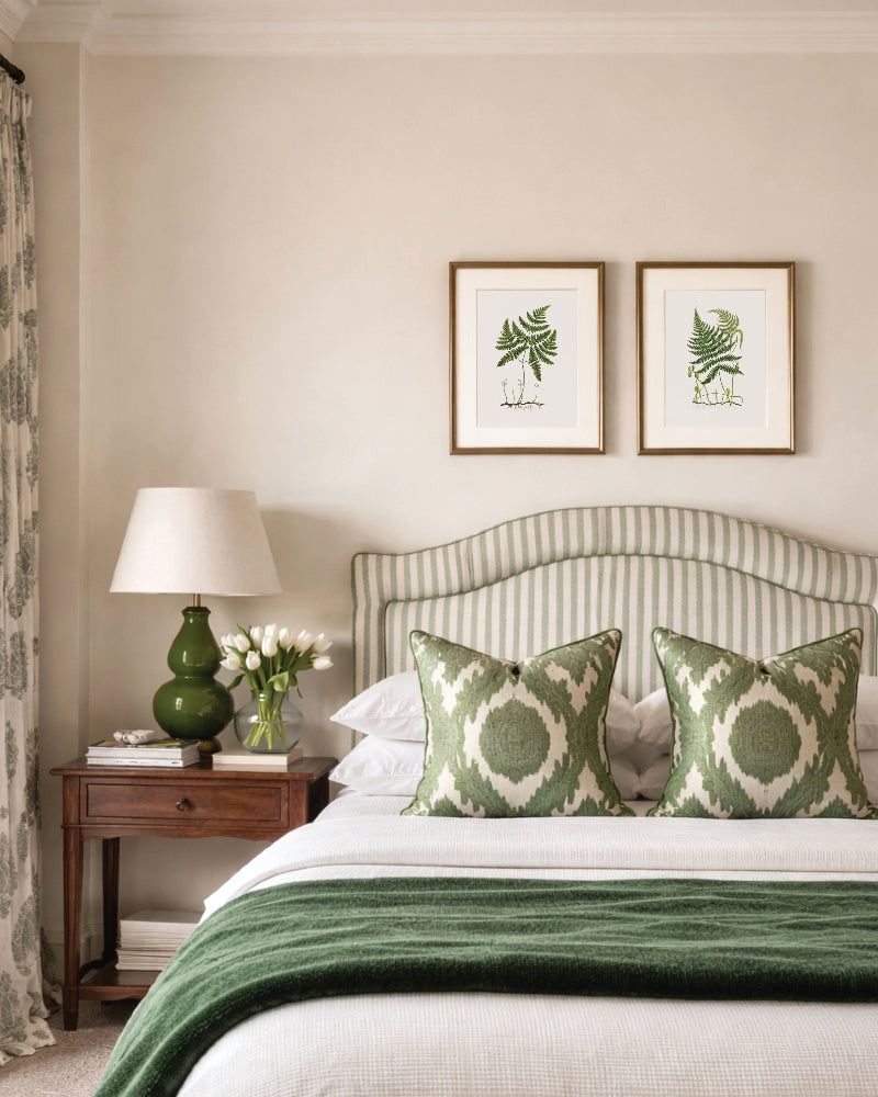 Neatly made bed with green and white patterned pillows, nightstand with lamp, and vintage wall art in a bedroom.