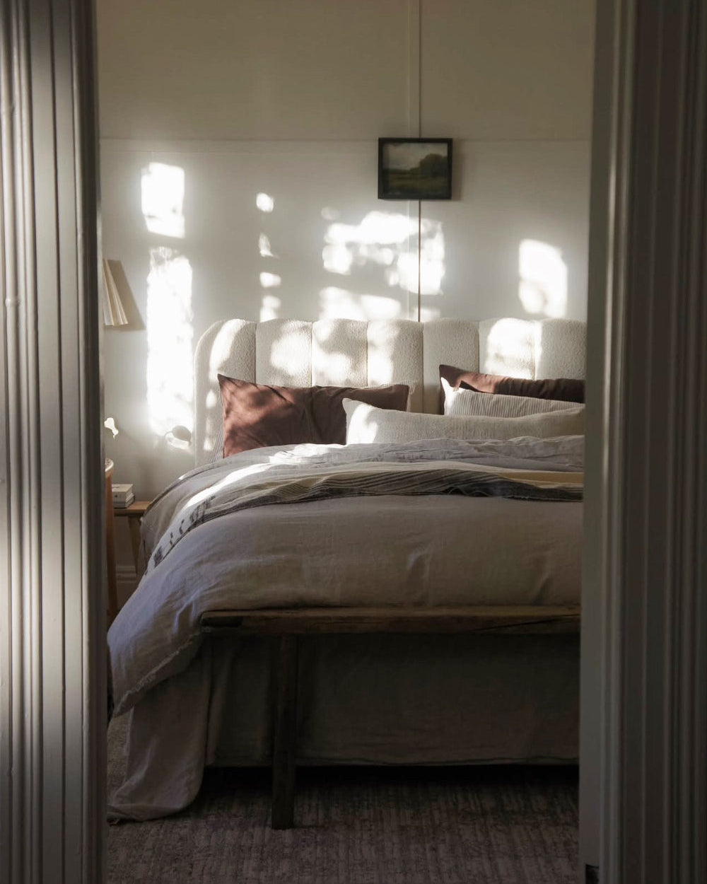 A softly lit bedroom scene viewed through an open doorway, featuring a bed dressed in light neutral bedding with dark brown pillows, a small wall art piece above, and dappled sunlight on the wall.