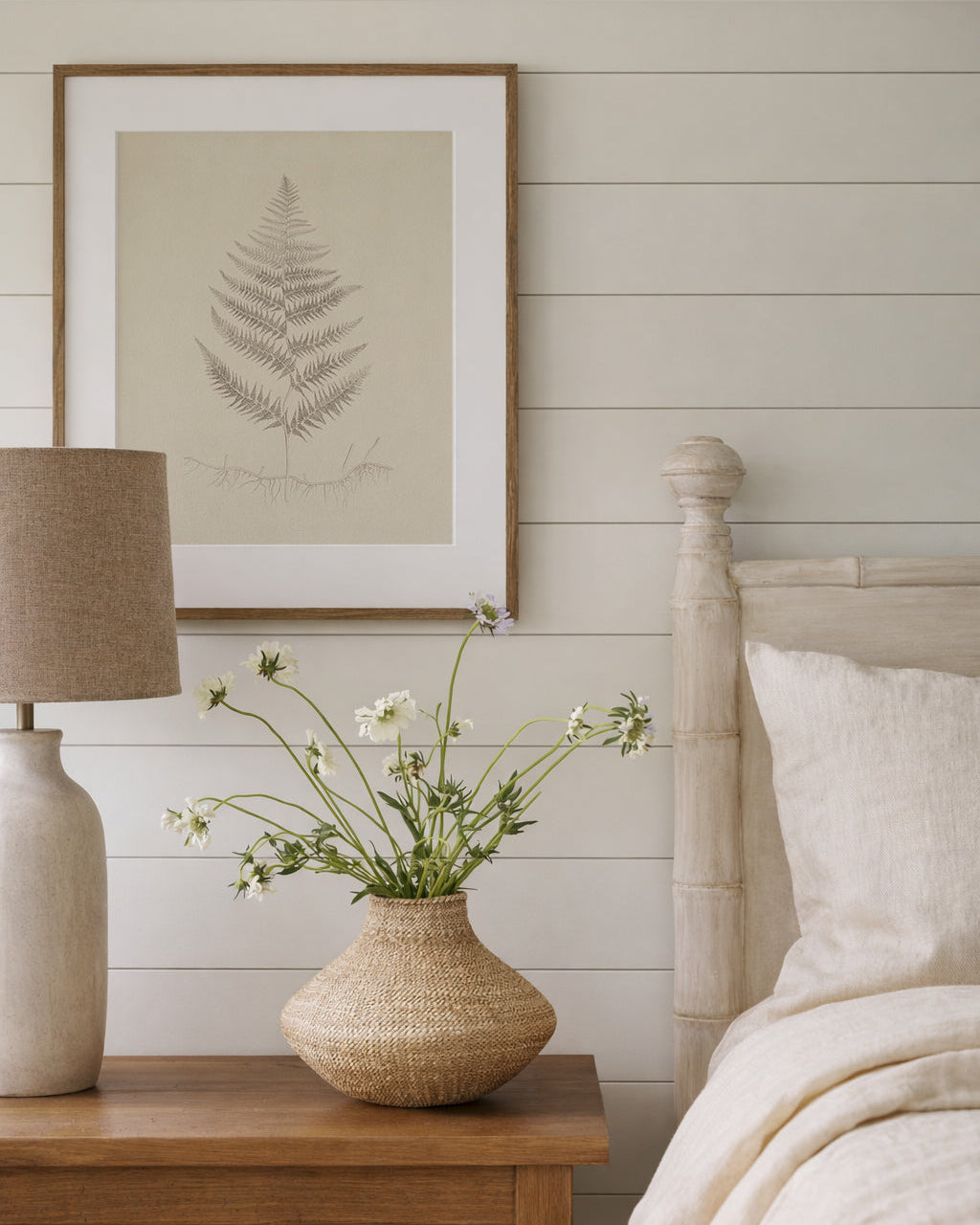 Bedroom with wooden nightstand, lamp, and framed fern artwork on a white wall.