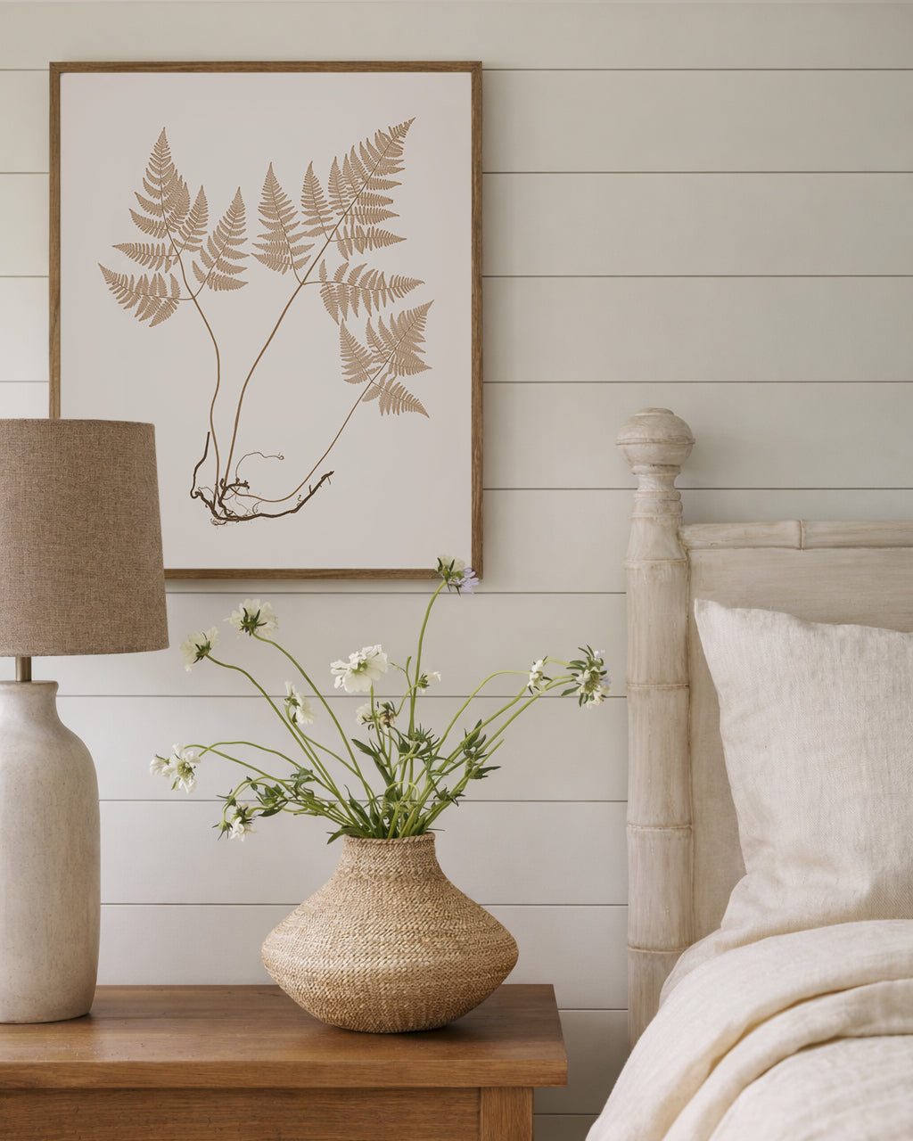 Stylish neutral Bedroom with wooden nightstand, lamp, and framed vintage botanical art on a white wall.