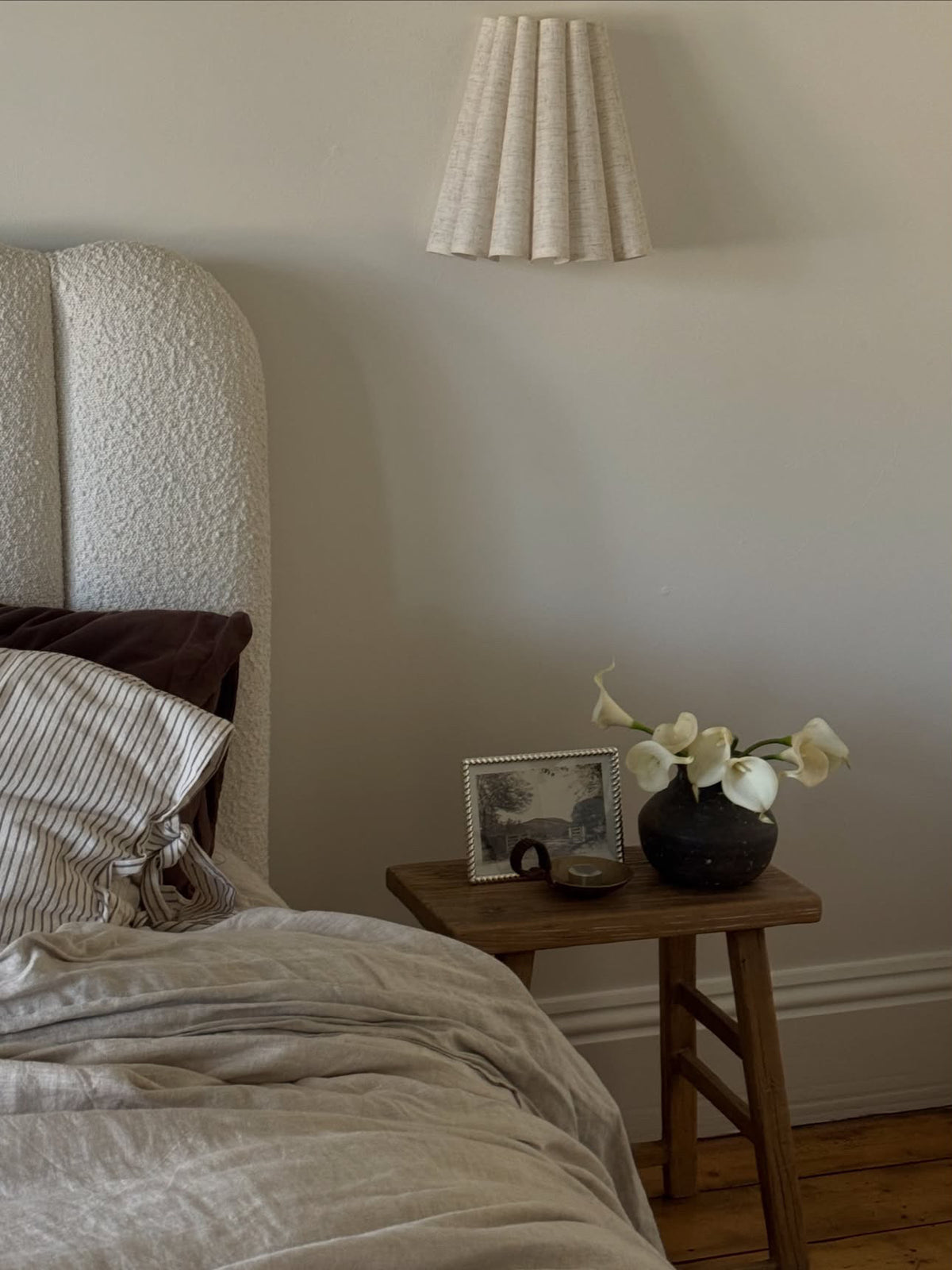 A cozy bedroom corner featuring a textured cream upholstered headboard, neutral beige bedding, a small wooden nightstand with a black vase of white flowers, and a matching pleated wall lamp on a beige wall.