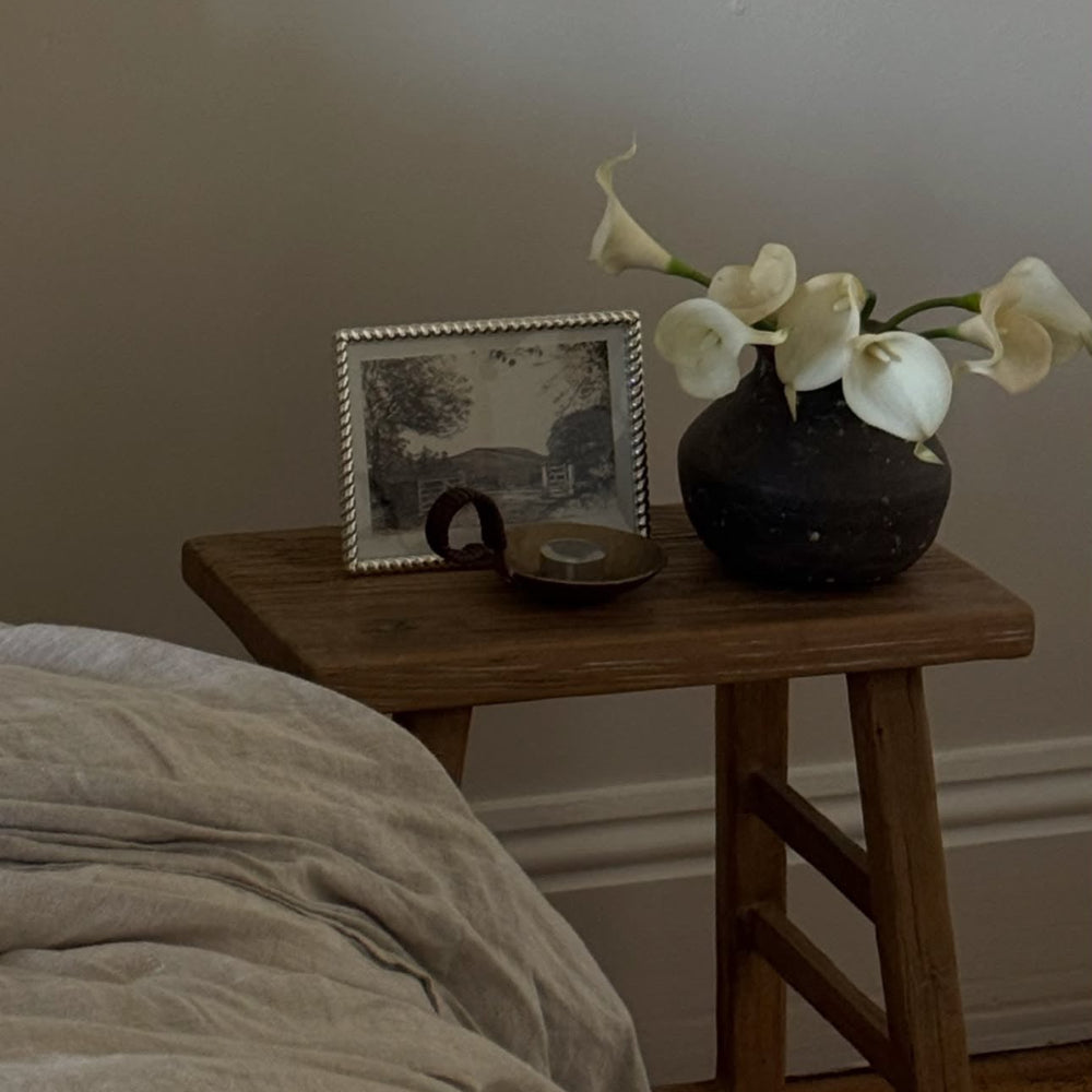 A rustic wooden nightstand beside a bed, styled with a small framed black-and-white photo, a dark ceramic candle holder, and a dark vase holding white calla lilies against a neutral wall.
